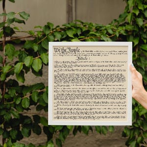 May include: A framed print of the US Constitution, with the text "We the People" at the top. The document is in black ink on a cream-colored background. The frame is white.