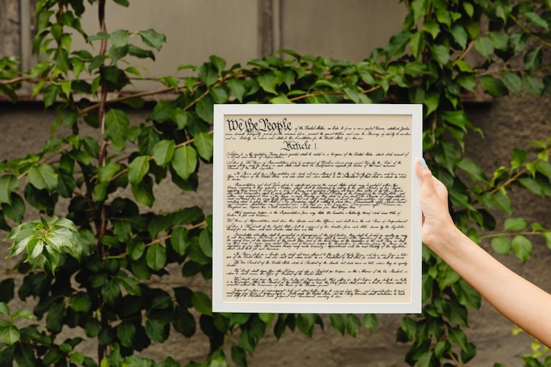 May include: A framed print of the US Constitution, with the text "We the People" at the top. The document is in black ink on a cream-colored background. The frame is white.