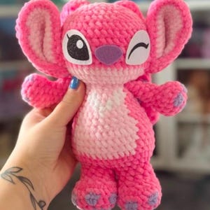 Può includere: Peluche rosa lavorato a uncinetto con un viso che strizza l'occhio e grandi orecchie. Il giocattolo è realizzato con un filato rosa e bianco e ha accenti viola sulle orecchie e sui piedi.