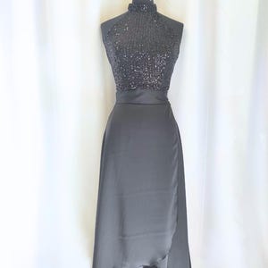May include: A black evening dress on a mannequin. The bodice is covered in sequins, and the skirt is a flowing, wrap-style design. The dress has a high neckline and a defined waist. The fabric appears to be a satin or similar material.