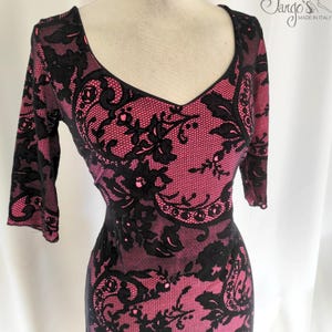 Puede incluir: Un vestido de encaje negro y rosa con escote en V y mangas 3/4. El vestido tiene un patrón floral y está hecho en Italia.