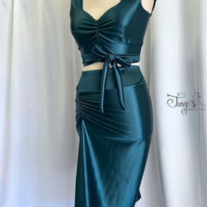 May include: A teal two-piece dance outfit on a mannequin. The top has a ruched front and a tie at the waist. The skirt is a midi length with a side slit. The fabric has a satin sheen. The outfit is from Tango's, Made in Italy.