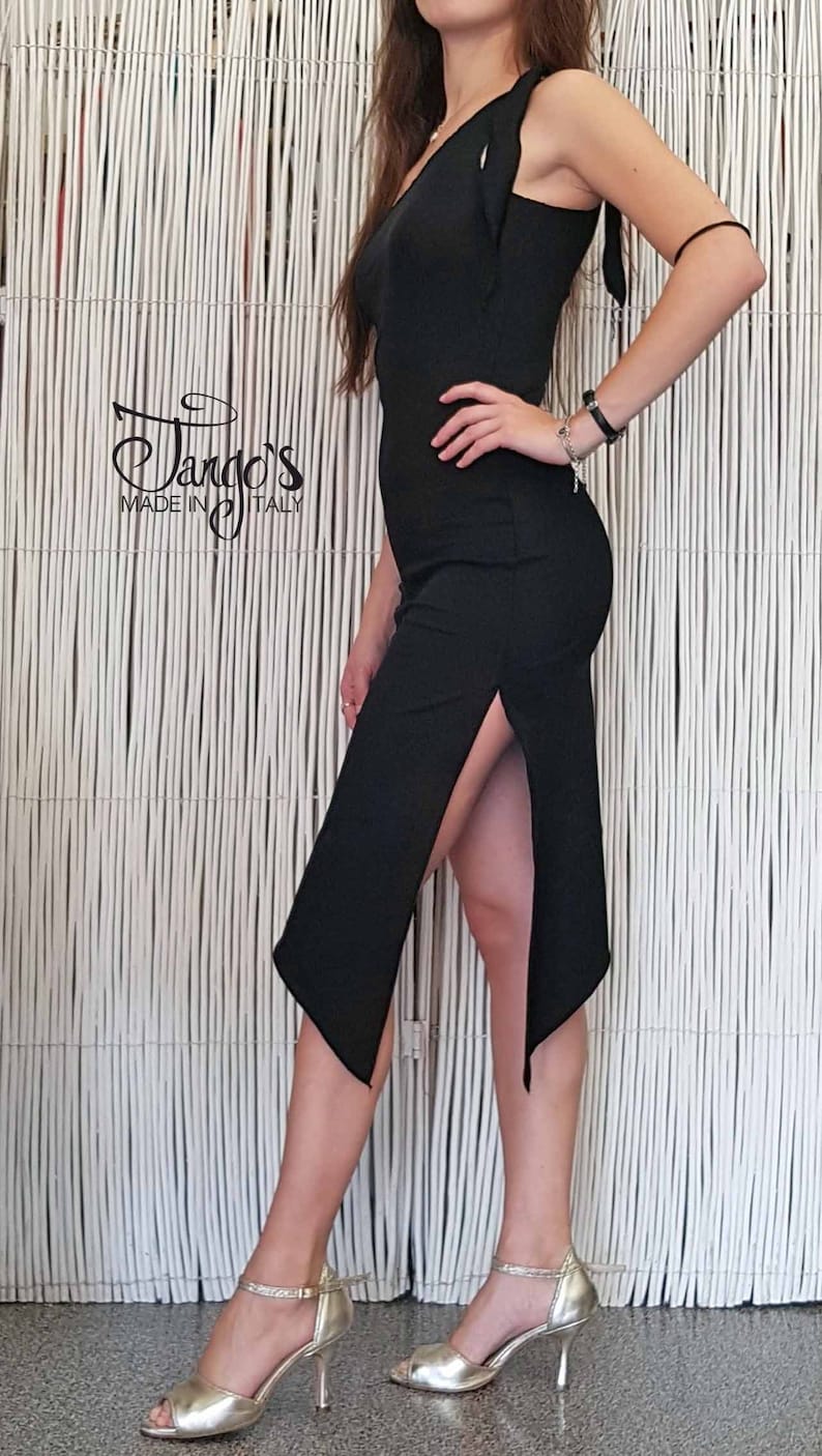 May include: A woman wearing a black dress with a side slit and a gold pair of heels. The dress has a strap that goes over the shoulder and a small ruffle detail. The woman is standing in front of a white slatted wall. The text "Tango's Made in Italy" is visible in the lower left corner of the image.