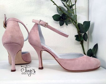 May include: A pair of light pink suede heeled shoes with ankle straps and a small rhinestone embellishment on the strap. The shoes are on a white surface with a green plant in the background. The text "Tango's Made in Italy" is visible on the surface.