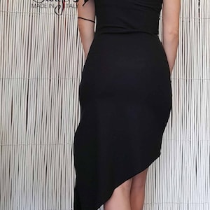 May include: A black one-shoulder dress with a ruffled detail on the shoulder. The dress has a high-low hemline and is made of a stretchy fabric. The model is wearing the dress with gold heels.