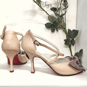 May include: A pair of beige leather dance shoes with a snakeskin bow detail. The shoes have a red heel and a strap across the ankle. The shoes are made in Italy by Tango's.