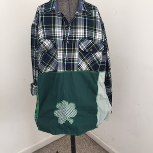 May include: A patchwork shirt on a dressmaker's dummy. The top half is a green, navy, and white plaid flannel. The bottom half is a solid green fabric with a four-leaf clover applique. The shirt has two chest pockets and a button-down front.