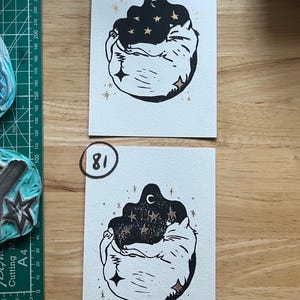 May include: Two rectangular white paper prints featuring a black ink linocut illustration of a curled-up cat surrounded by gold foil stars and a crescent moon. The top print is labeled 80 and the bottom print is labeled 81 in black ink.