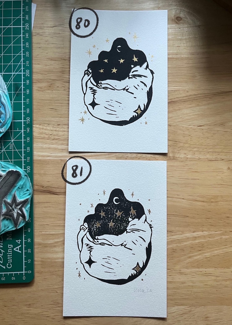 May include: Two rectangular white paper prints featuring a black ink linocut illustration of a curled-up cat surrounded by gold foil stars and a crescent moon. The top print is labeled 80 and the bottom print is labeled 81 in black ink.