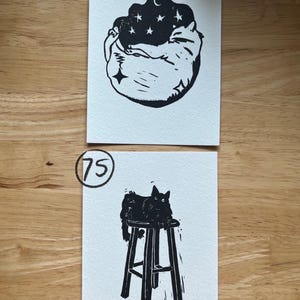 May include: Two rectangular white paper prints with black ink illustrations. The top print, labeled 74, shows a curled-up cat beneath a dark sky with stars and a crescent moon. The bottom print, labeled 75, depicts a cat sitting on a tall stool.
