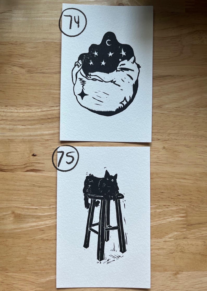 May include: Two rectangular white paper prints with black ink illustrations. The top print, labeled 74, shows a curled-up cat beneath a dark sky with stars and a crescent moon. The bottom print, labeled 75, depicts a cat sitting on a tall stool.