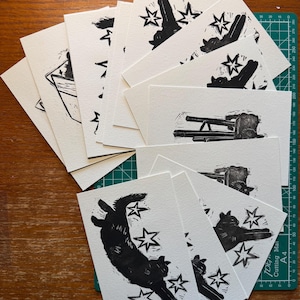 Pu&ograve; includere: Una collezione di cartoline rettangolari in carta bianca con stampe a inchiostro nero. I disegni includono sagome di gatti in varie pose, alcuni circondati da forme a stella e altri raffigurati mentre riposano su mobili o in scatole.
