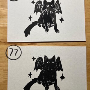 May include: Two rectangular white cards featuring black ink illustrations of a cat with bat wings. The top card is labeled with the number 76 in a circle, and the bottom card is labeled with the number 77 in a circle. Both show a cat with four-pointed stars.