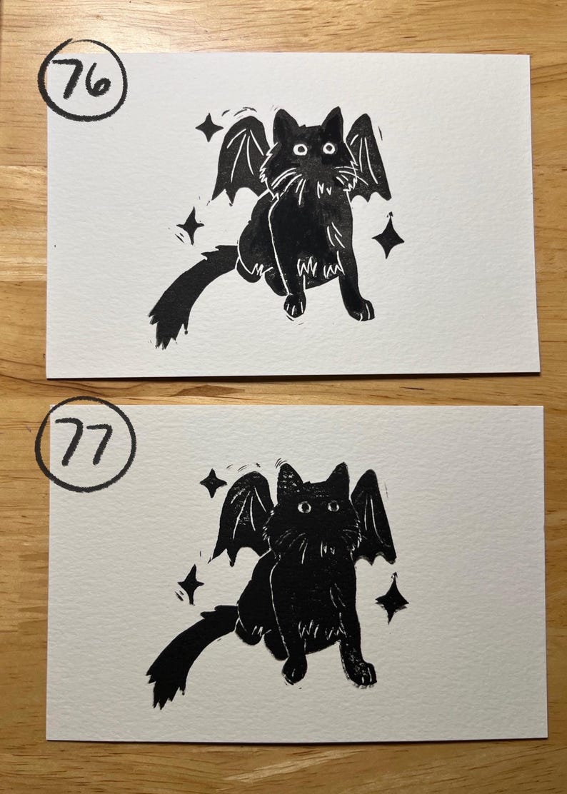 May include: Two rectangular white cards featuring black ink illustrations of a cat with bat wings. The top card is labeled with the number 76 in a circle, and the bottom card is labeled with the number 77 in a circle. Both show a cat with four-pointed stars.