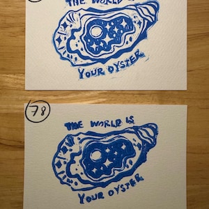 May include: Two rectangular white cards featuring a blue ink block print of an oyster shell containing a moon and stars. The text "THE WORLD IS YOUR OYSTER" is printed in blue. The top card is marked with the number 18 and the bottom with 78.
