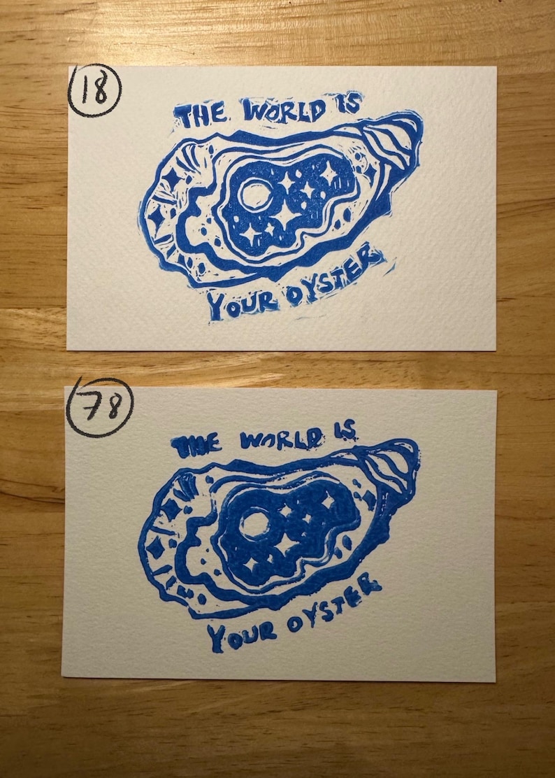 May include: Two rectangular white cards featuring a blue ink block print of an oyster shell containing a moon and stars. The text "THE WORLD IS YOUR OYSTER" is printed in blue. The top card is marked with the number 18 and the bottom with 78.