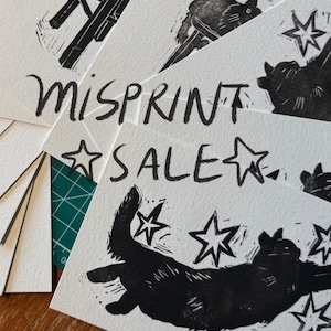 Pu&ograve; includere: Una pila di cartoline rettangolari bianche con stampe a inchiostro nero di un gatto che cerca di afferrare delle stelle. Il testo MISPRINT SALE &egrave; stampato in un carattere nero disegnato a mano sulle carte, con illustrazioni di stelle.