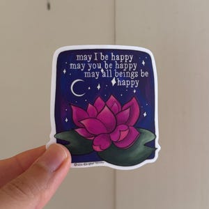May include: A sticker with a pink lotus flower on a dark blue background with white stars and a crescent moon. The text reads "may I be happy may you be happy may all beings be happy".