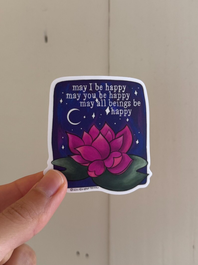May include: A sticker with a pink lotus flower on a dark blue background with white stars and a crescent moon. The text reads "may I be happy may you be happy may all beings be happy".