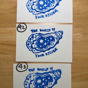 Pu&ograve; includere: Tre cartoline rettangolari bianche con una stampa a inchiostro blu di un guscio di ostrica. All'interno del guscio c'&egrave; un motivo con luna e stelle. Il testo "THE WORLD IS YOUR OYSTER" &egrave; stampato in inchiostro blu. Ogni cartolina &egrave; contrassegnata da un numero cerchiato: 18, 42 o 43.