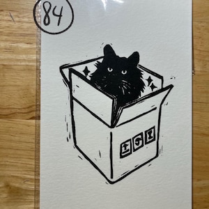 May include: A black and white linocut print on paper featuring a fluffy black cat sitting inside an open cardboard box. The box has three small square icons on the front and the number 84 is circled in the top left corner of the white paper.