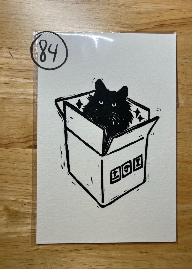 May include: A black and white linocut print on paper featuring a fluffy black cat sitting inside an open cardboard box. The box has three small square icons on the front and the number 84 is circled in the top left corner of the white paper.