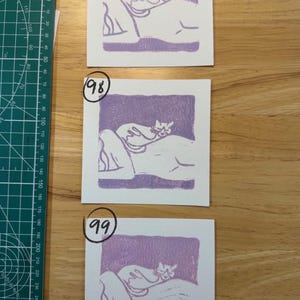 May include: Three square white paper prints featuring a purple block-printed illustration of a cat resting on a person's back. Each print is labelled with a hand-written number 97, 98, or 99. A green cutting mat with metric markings is visible.
