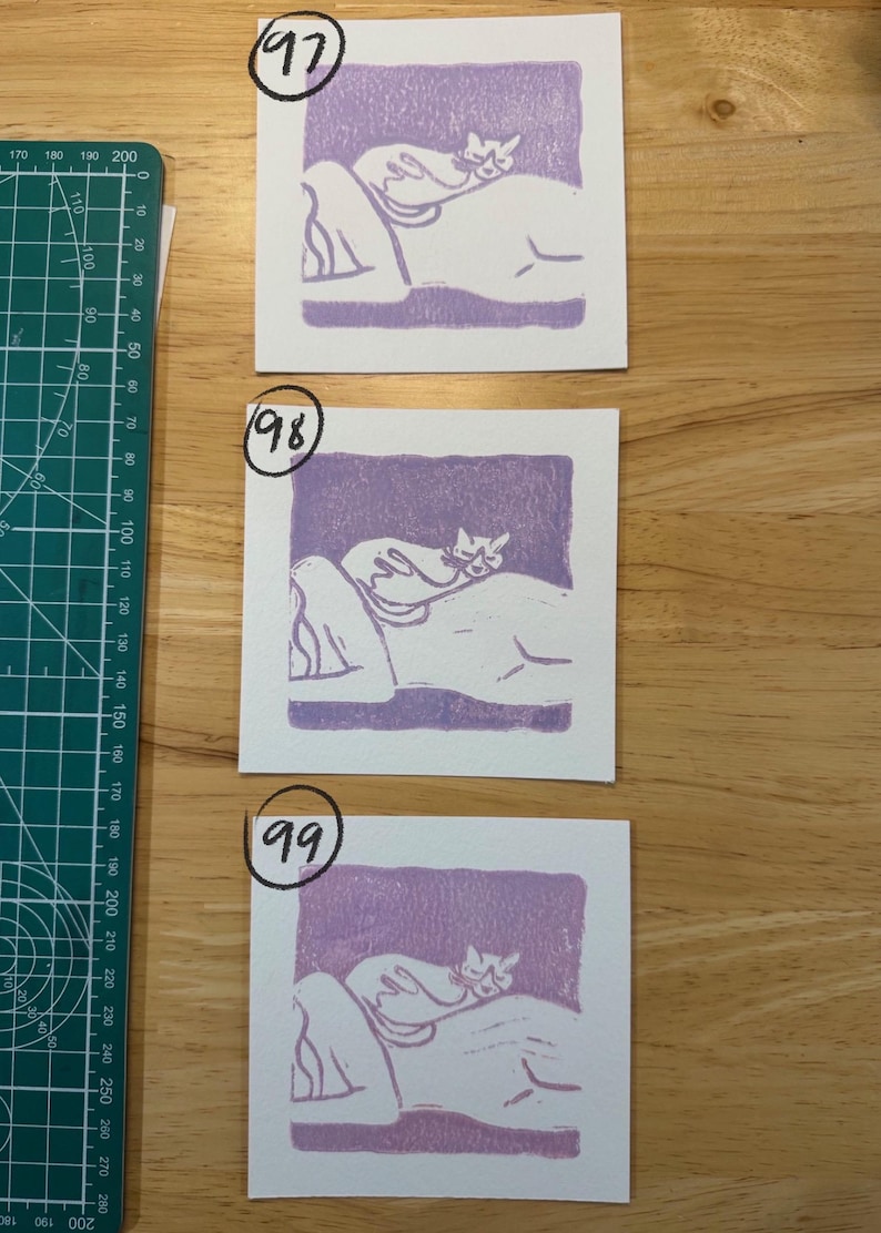 May include: Three square white paper prints featuring a purple block-printed illustration of a cat resting on a person's back. Each print is labelled with a hand-written number 97, 98, or 99. A green cutting mat with metric markings is visible.