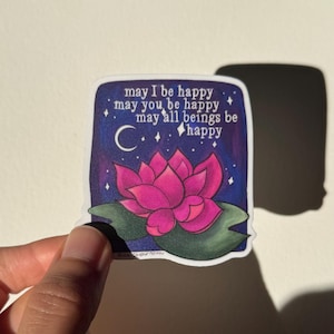 May include: A sticker with a pink lotus flower on a dark blue background with white stars and a crescent moon. The text reads "may I be happy, may you be happy, may all beings be happy".