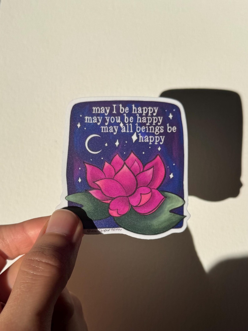 May include: A sticker with a pink lotus flower on a dark blue background with white stars and a crescent moon. The text reads "may I be happy, may you be happy, may all beings be happy".