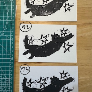 May include: Three rectangular white paper prints featuring a black ink block print of a cat reaching toward four stars. Each print is labelled with a circled number: 91, 92, and 93. A green cutting mat with metric markings is visible on the left side.