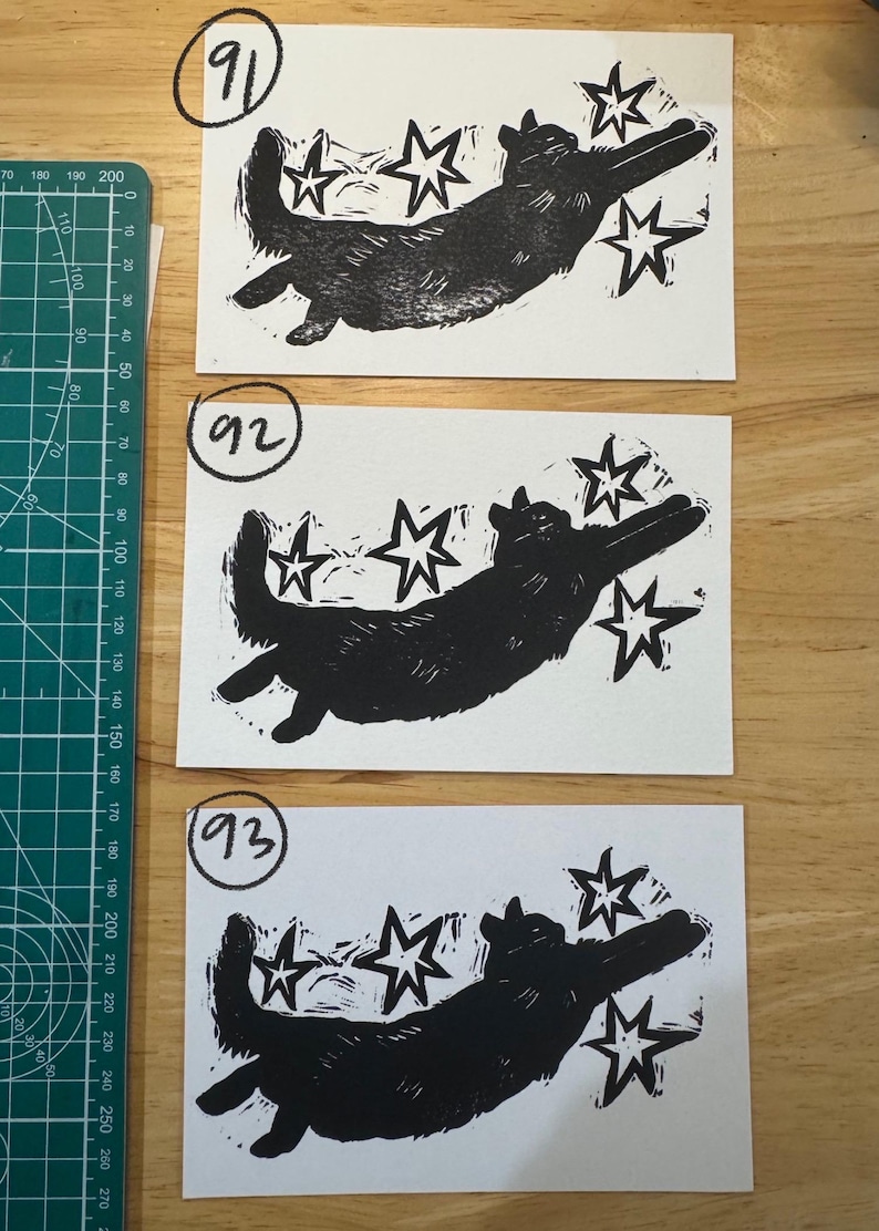 May include: Three rectangular white paper prints featuring a black ink block print of a cat reaching toward four stars. Each print is labelled with a circled number: 91, 92, and 93. A green cutting mat with metric markings is visible on the left side.