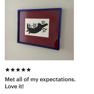 May include: Framed artwork showing a black cat silhouette with stars on a white background. The artwork has a burgundy mat and a blue frame, mounted on a white wall. A customer review is visible below the artwork.