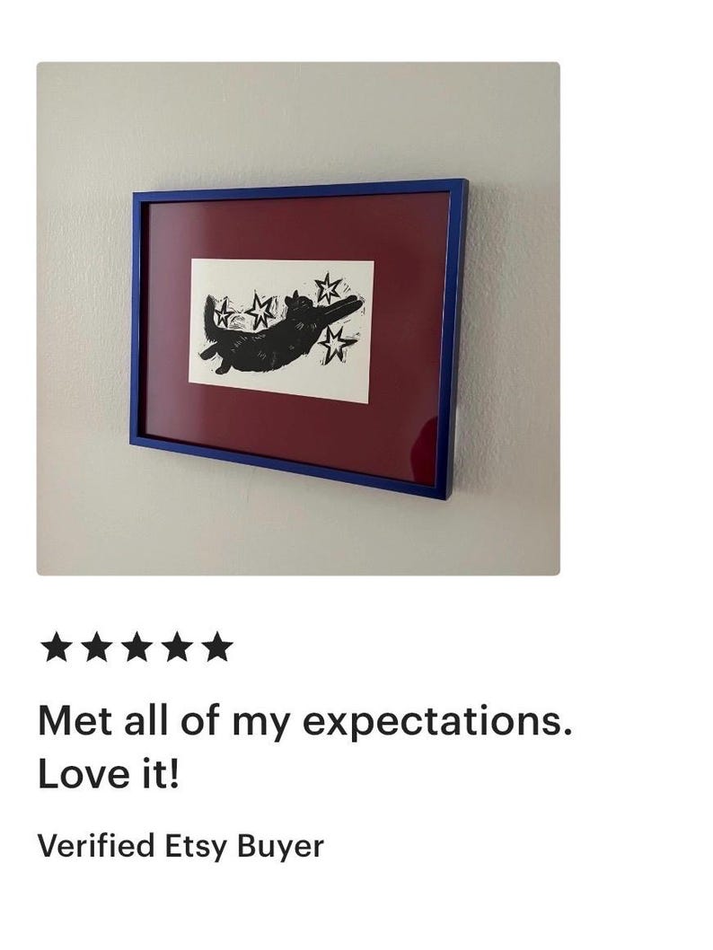 May include: Framed artwork showing a black cat silhouette with stars on a white background. The artwork has a burgundy mat and a blue frame, mounted on a white wall. A customer review is visible below the artwork.