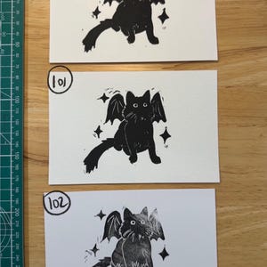 May include: Three rectangular white cards featuring black ink block prints of a winged cat surrounded by four-pointed stars. Each card is labelled with a hand-drawn number 100, 101, or 102 in a circle. A green cutting mat with metric markings is visible.