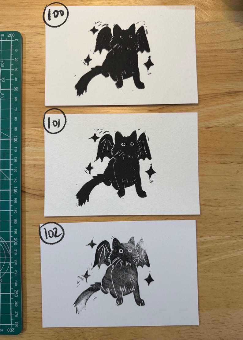 May include: Three rectangular white cards featuring black ink block prints of a winged cat surrounded by four-pointed stars. Each card is labelled with a hand-drawn number 100, 101, or 102 in a circle. A green cutting mat with metric markings is visible.