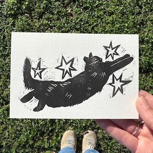 May include: A black and white linocut print of a dog with stars. The dog is depicted in a playful pose, with its legs extended. The print is on a white rectangular piece of paper.
