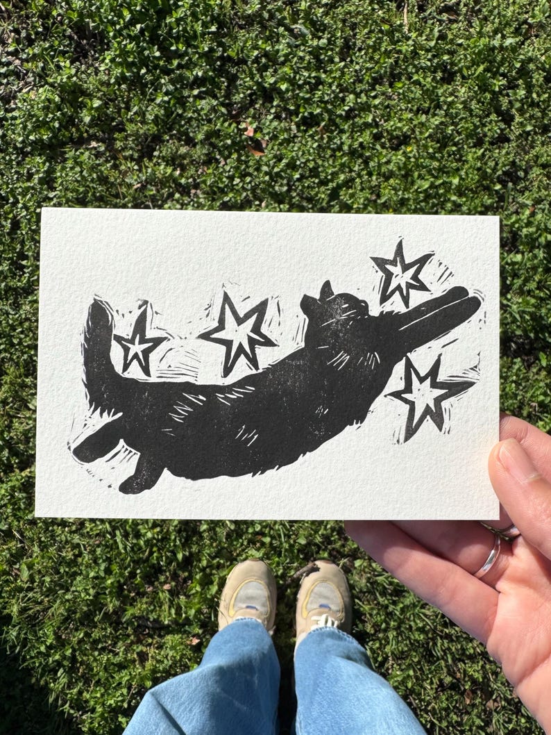 May include: A black and white linocut print of a dog with stars. The dog is depicted in a playful pose, with its legs extended. The print is on a white rectangular piece of paper.