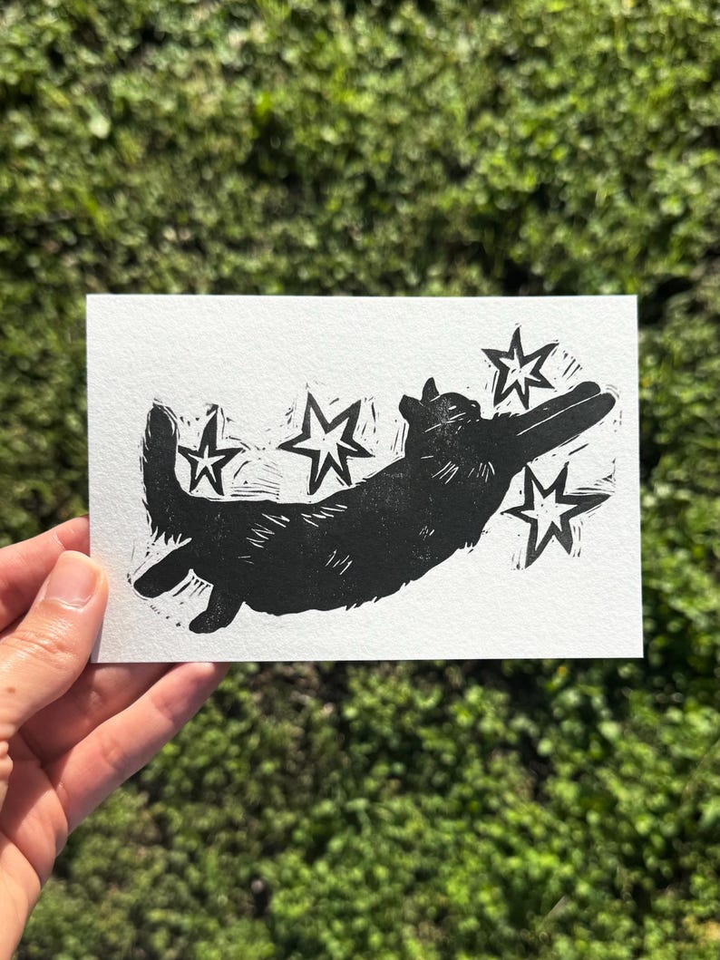 May include: A black and white linocut print of a corgi dog leaping amongst stars. The artwork is on a white rectangular card, held against a blurred green background. The dog and stars are solid black.
