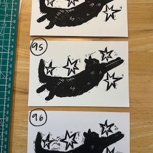 May include: Three rectangular white paper prints featuring a black ink block print of a cat reaching towards four stars. Each print is labelled with a handwritten number 94, 95, or 96. A green ruler is visible on the left, showing a length of 28 cm.