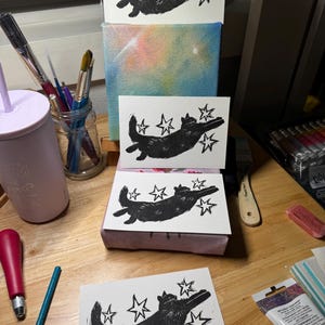 May include: A collection of black and white linocut prints featuring a cat design with stars. The cat is shown in a playful pose, with star shapes around it. The prints are arranged on a wooden surface, with art supplies such as paintbrushes and a canvas with a colourful background.