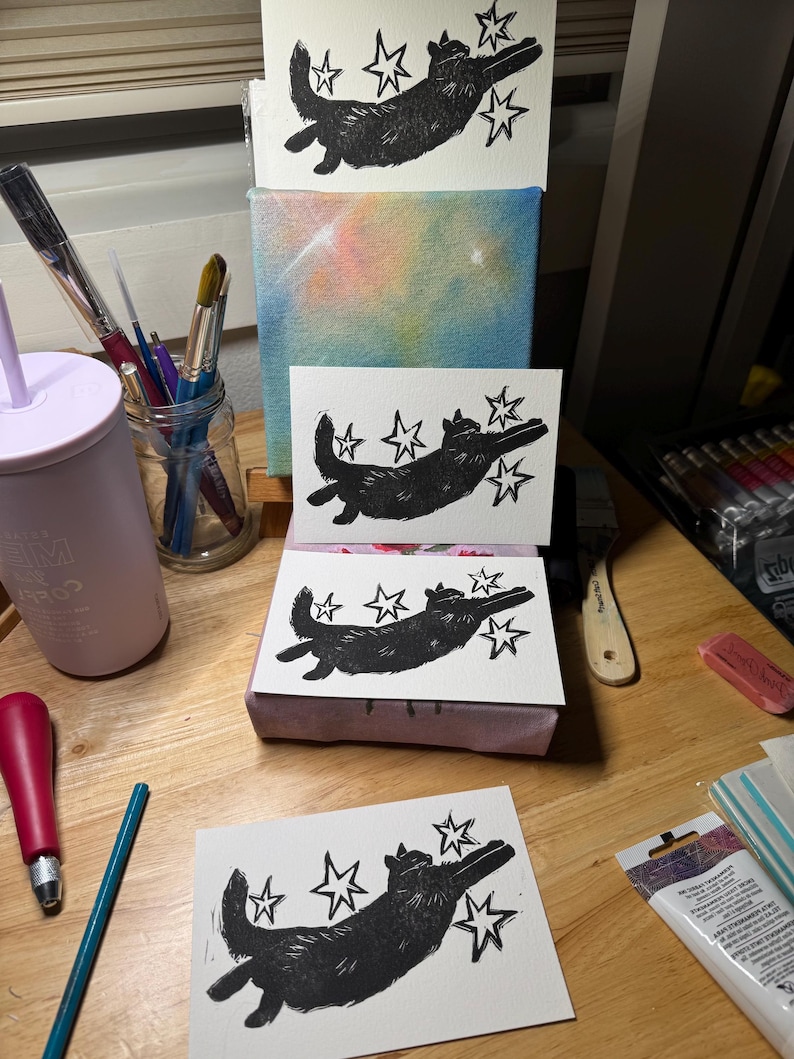 May include: A collection of black and white linocut prints featuring a cat design with stars. The cat is shown in a playful pose, with star shapes around it. The prints are arranged on a wooden surface, with art supplies such as paintbrushes and a canvas with a colourful background.