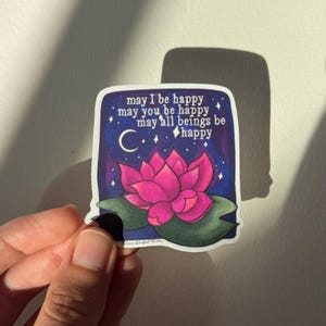 May include: A sticker with a pink lotus flower on a dark blue background with white stars and a crescent moon. The text reads "may I be happy may you be happy may all beings be happy".