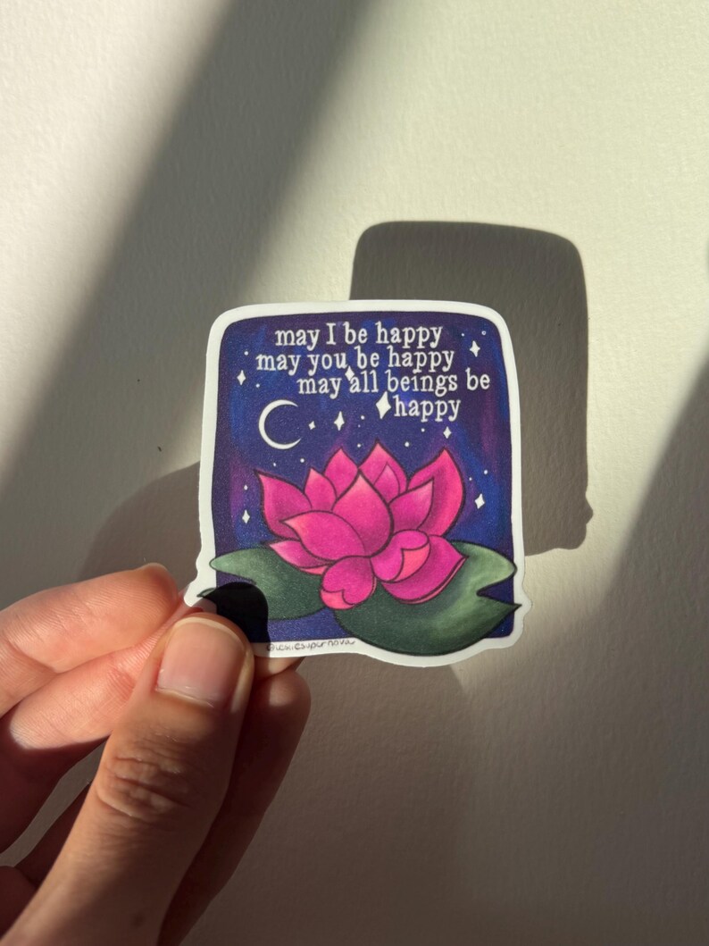 May include: A sticker with a pink lotus flower on a dark blue background with white stars and a crescent moon. The text reads "may I be happy may you be happy may all beings be happy".