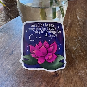 May include: A sticker with a pink lotus flower on a dark blue background with white stars and a crescent moon. The text reads "may I be happy, may you be happy, may all beings be happy".