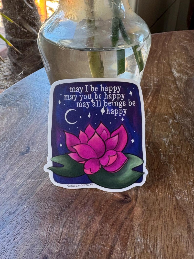 May include: A sticker with a pink lotus flower on a dark blue background with white stars and a crescent moon. The text reads "may I be happy, may you be happy, may all beings be happy".