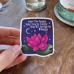 May include: A sticker with a pink lotus flower on a dark blue background with white stars and a crescent moon. The sticker has the text "may I be happy, may you be happy, may all beings be happy" and the signature "Gucn.csupernova".
