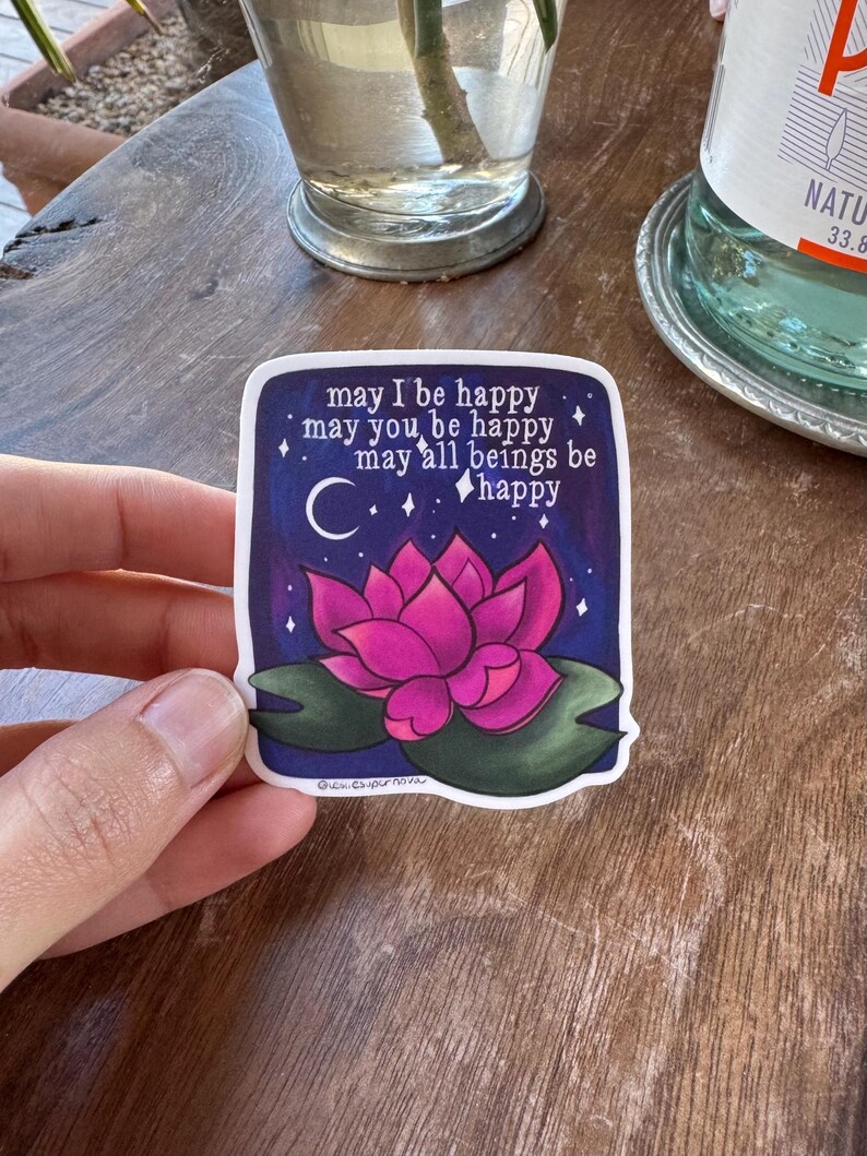 May include: A sticker with a pink lotus flower on a dark blue background with white stars and a crescent moon. The sticker has the text "may I be happy, may you be happy, may all beings be happy" and the signature "Gucn.csupernova".