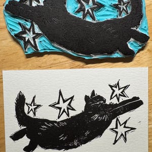 May include: A black cat and star linocut print and its corresponding stamp. The stamp is black with a light blue background. The print shows a cat with outstretched paws and stars.