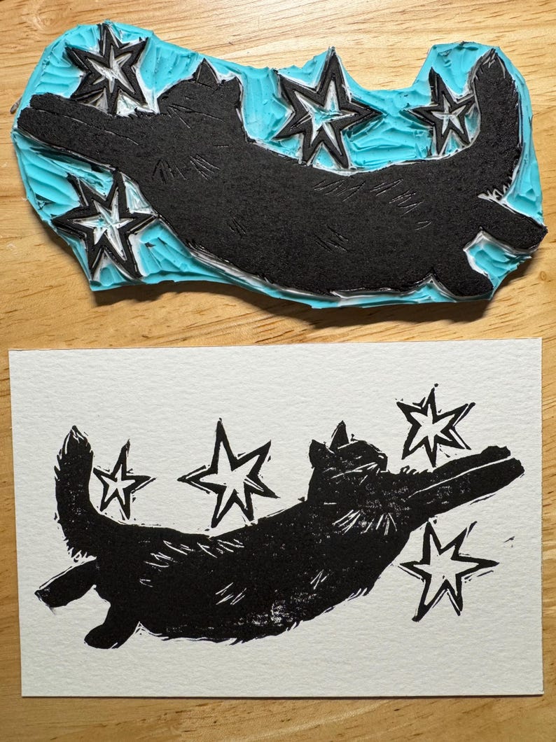 May include: A black cat and star linocut print and its corresponding stamp. The stamp is black with a light blue background. The print shows a cat with outstretched paws and stars.